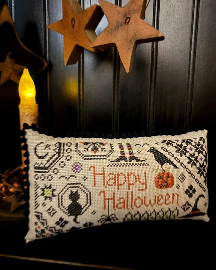 A rectangular cross-stitch pillow with a black and white pattern featuring the words 'Happy Halloween' in orange, a black cat silhouette, a pumpkin, and a crow, displayed against a dark paneled background with wooden star decorations and a lit candle.