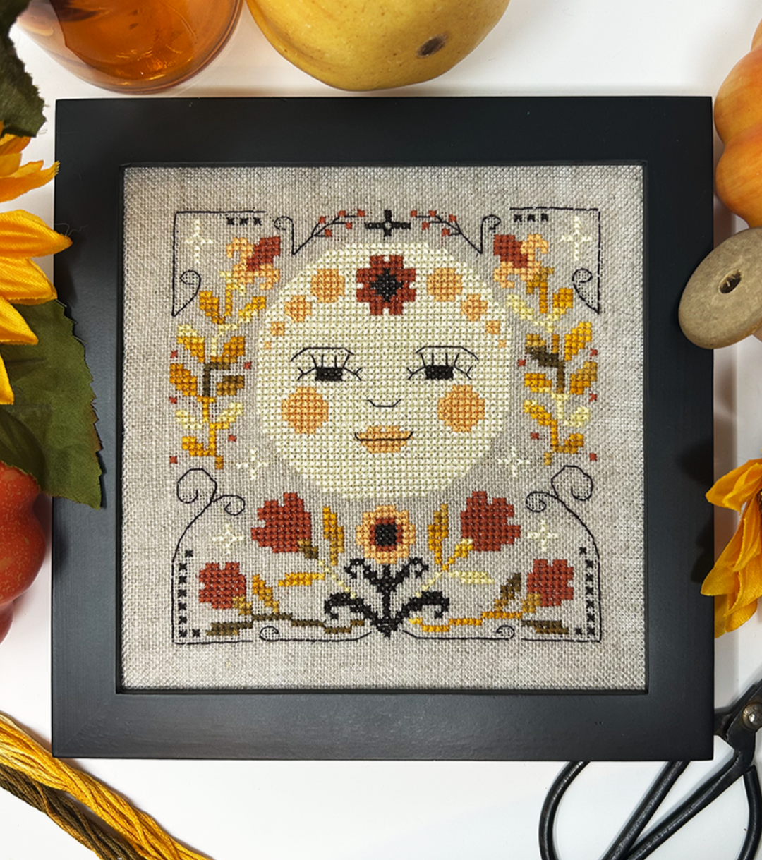 Framed cross-stitched artwork of a face with floral patterns, surrounded by autumn-themed items.