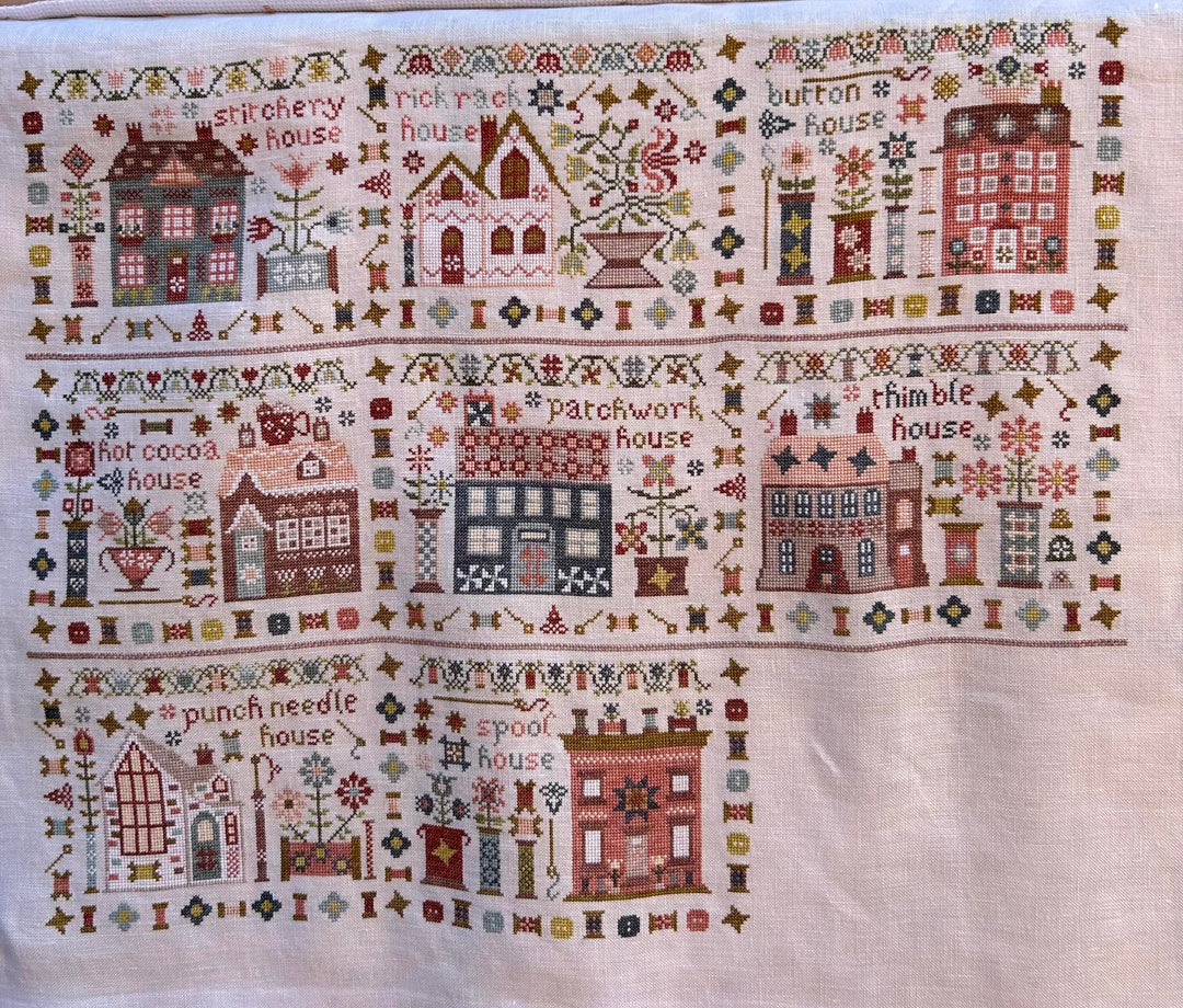 A square cross-stitch sampler tapestry featuring whimsical house illustrations and embroidered text such as 'patchwork house,' 'thimble house,' and 'punch needle house' on a cream fabric background.