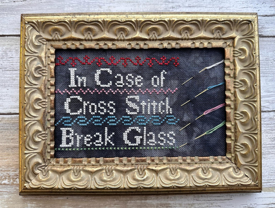 Decorative framed cross-stitched text 'In Case of Cross Stitch Break Glass' on a wooden surface.