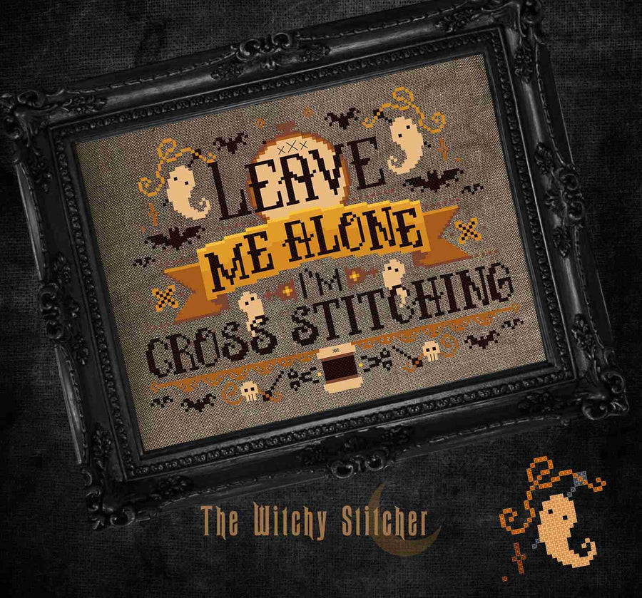 A framed cross-stitch artwork with a Halloween theme and the text 'Leave Me Alone I'm Cross Stitching.' The design includes ghosts, bats, skulls, and a small pumpkin on a tan fabric background.