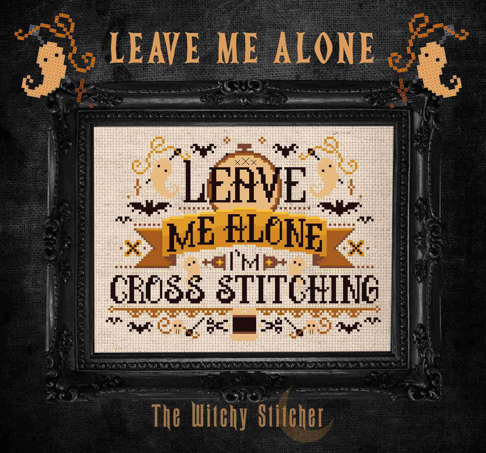 A framed cross-stitch design with the text 'LEAVE ME ALONE I'M CROSS STITCHING' in black and tan on a cream fabric background, surrounded by Halloween-themed motifs bats, ghosts, and spiders.