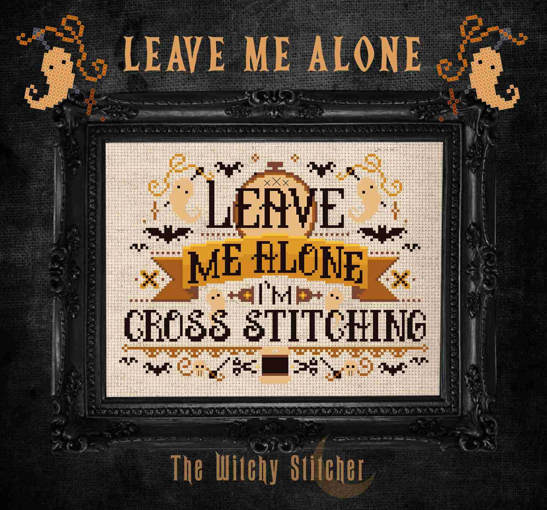 A framed cross-stitch design with the text 'LEAVE ME ALONE I'M CROSS STITCHING' in black and tan on a cream fabric background, surrounded by Halloween-themed motifs bats, ghosts, and spiders.