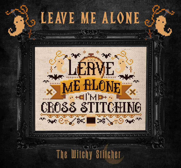 A framed cross-stitch design with the text 'LEAVE ME ALONE I'M CROSS STITCHING' in black and tan on a cream fabric background, surrounded by Halloween-themed motifs bats, ghosts, and spiders.