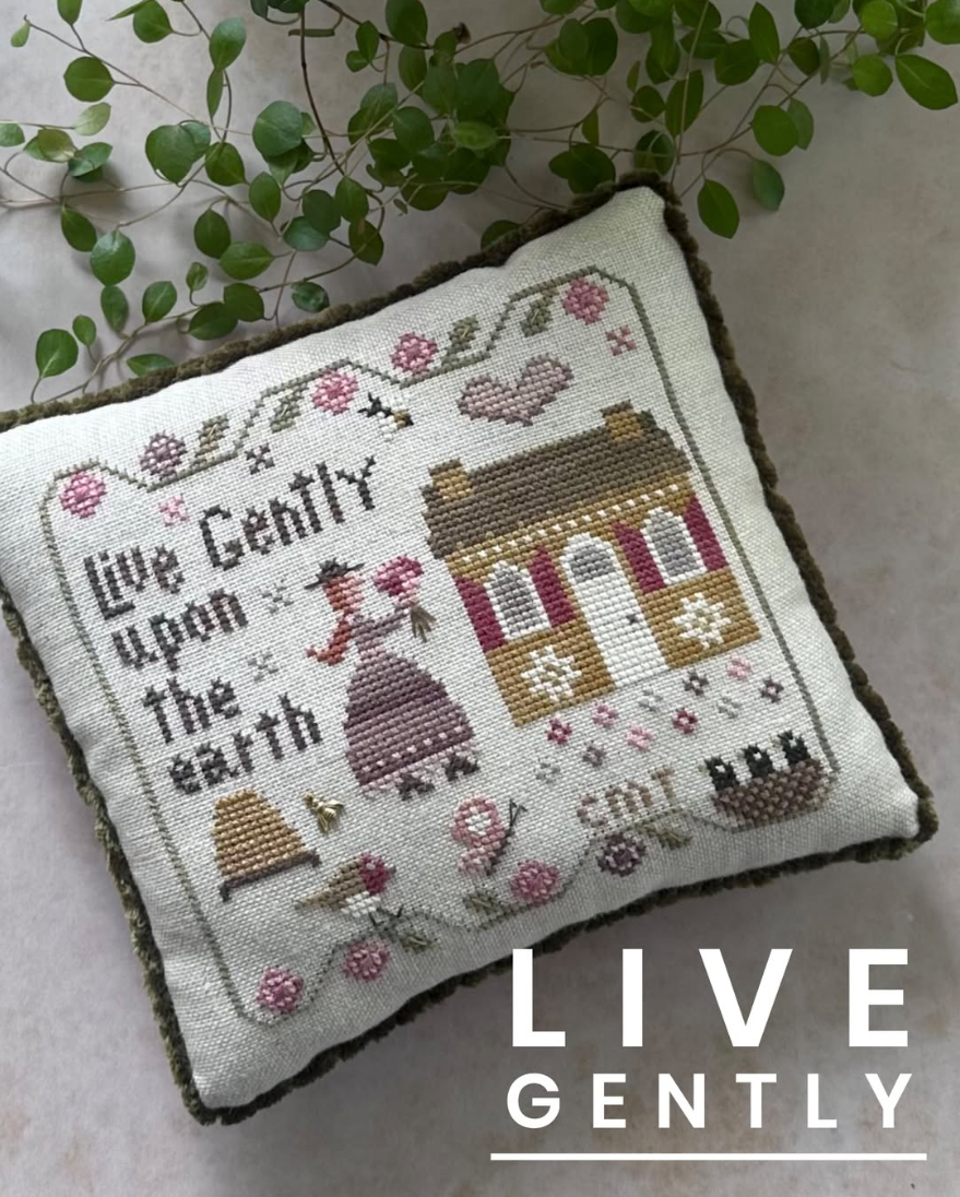 A square cross-stitch pillow with a rustic country design featuring a cottage, a woman, flowers, and the stitched text 'Live Gently upon the earth.' The pillow has a dark olive green border and is styled with green leafy vines on a light neutral background.