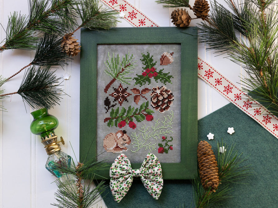 Framed cross-stitched design with Christmas elements, surrounded by pine branches and decorative items.