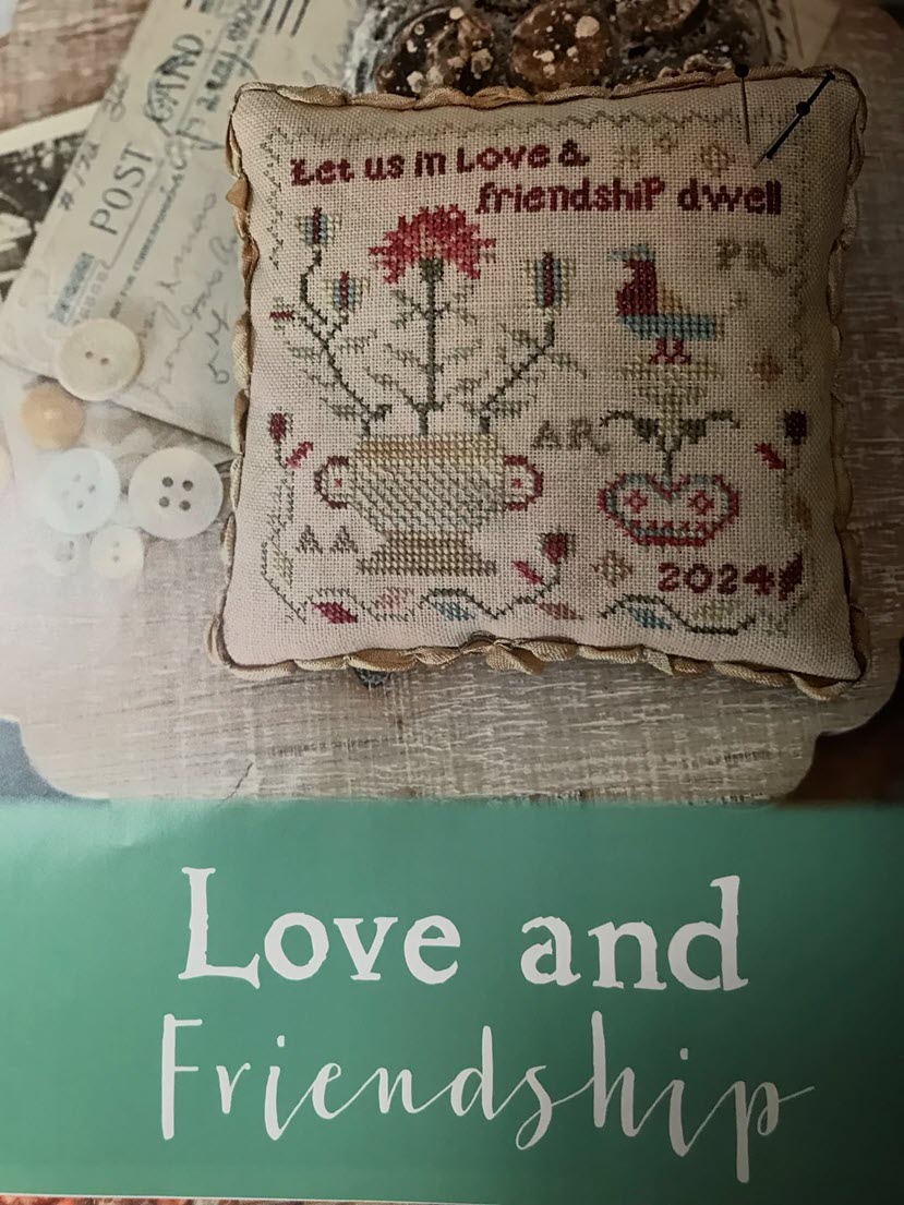 Decorative pillow with cross-stitch design and text 'Let us in love & friendship dwell' on a wooden surface.