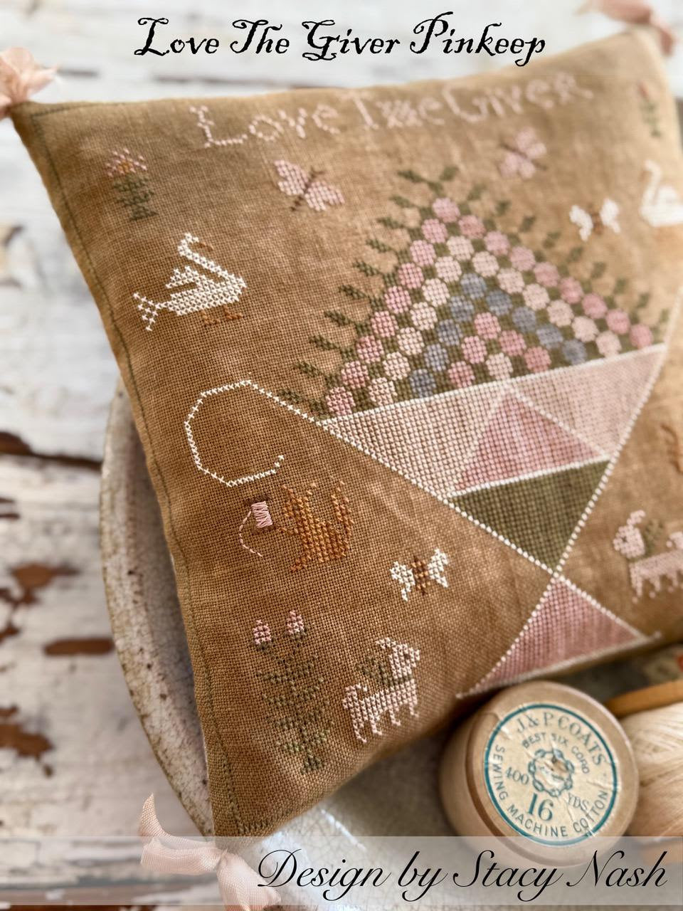 Embroidered pillow with cross-stitch patterns and 'Love The Giver Pinkeep' branding.