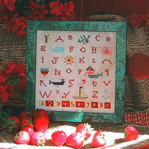 Framed cross-stitched alphabet and number chart with decorative elements on a table.