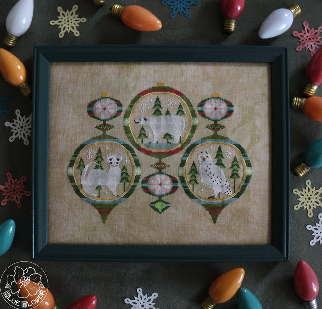 Framed cross-stitched artwork with winter-themed design on a decorative background with Christmas lights.