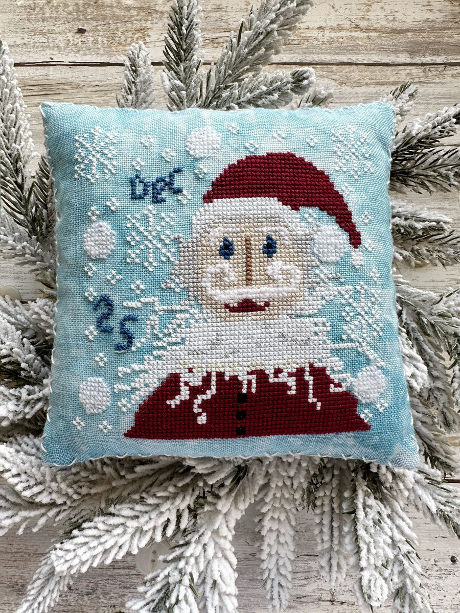 Cross-stitched Santa Claus pillow with snowflakes and 'Dec 25' text, placed on a wooden surface with pine branches.