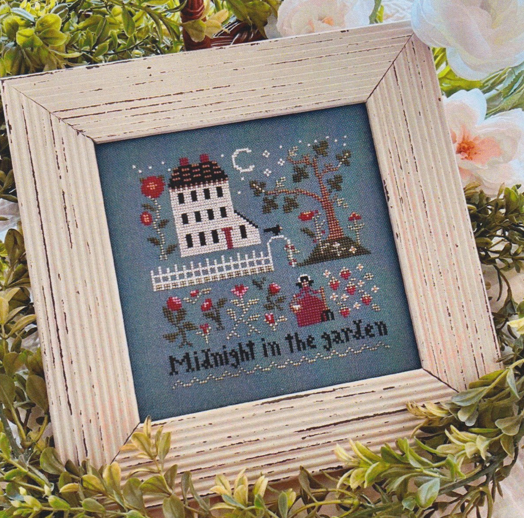 A framed needlework piece with a garden and moonlight scene, featuring a house, trees, and decorative elements in a blue background.
