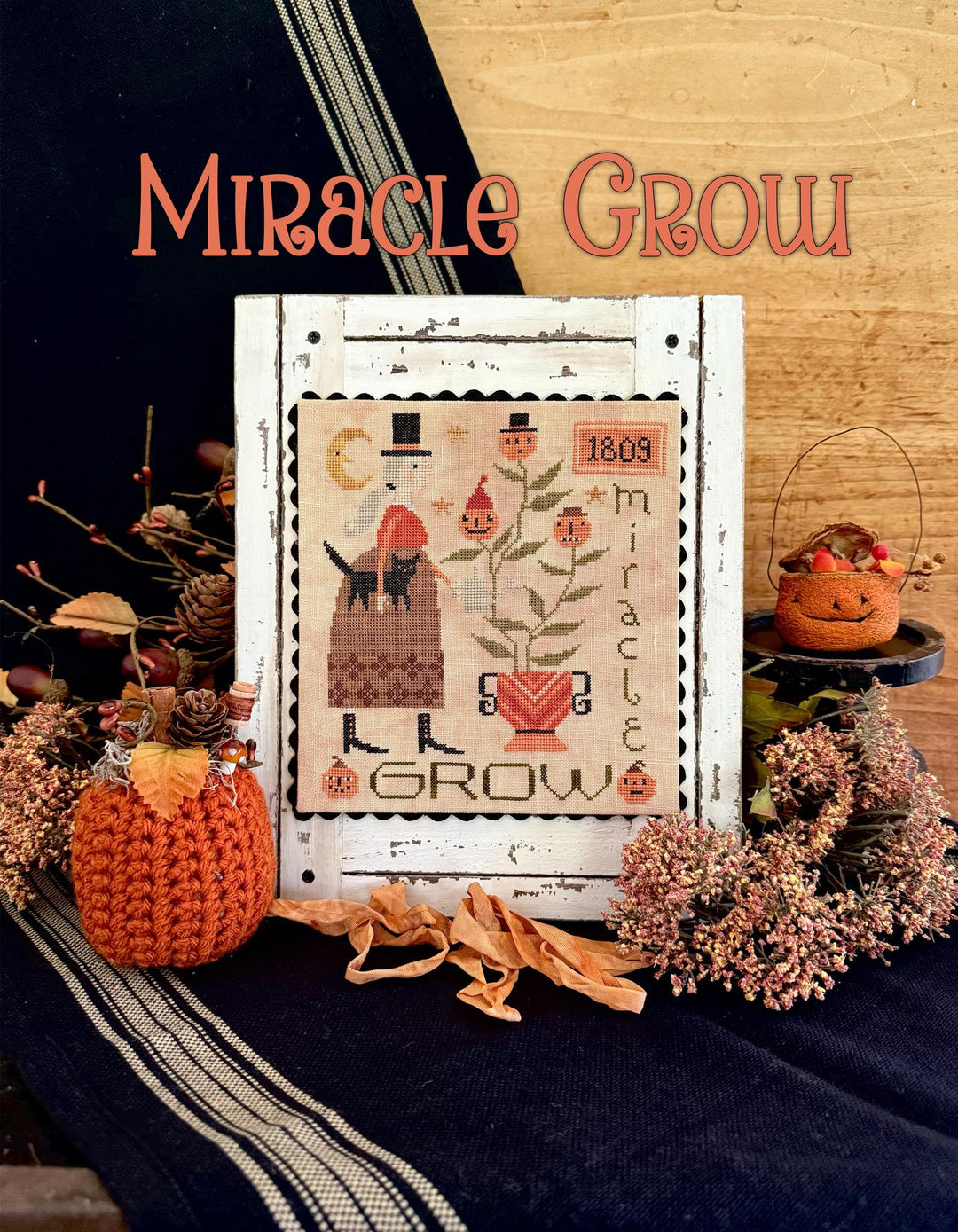 Decorative cross-stitch pattern with 'Miracle Grow' text on a wooden surface with pumpkins and flowers.