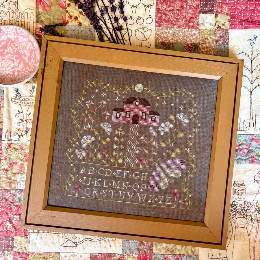 A framed cross-stitch sampler featuring a pink house, floral details, and an alphabet, displayed on a colorful patchwork quilt.
