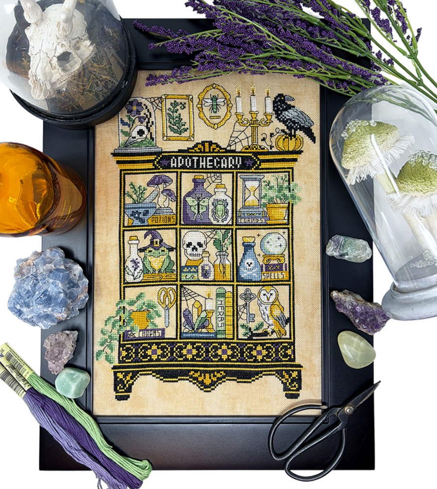 A framed witch-themed apothecary needlework or cross-stitch design displayed on a black board, surrounded by crystals, lavender, scissors, thread, and glass domes in a styled flat-lay arrangement.