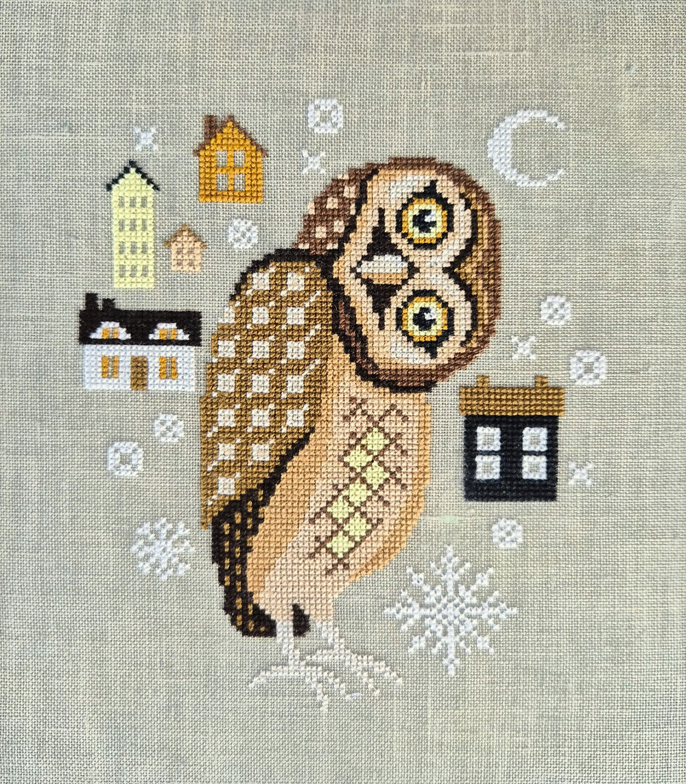 A cross-stitch needlework design on a light beige linen fabric, featuring a brown and tan owl surrounded by small houses, snowflakes, and a crescent moon.