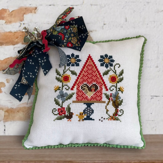 Decorative pillow with cross-stitched design of a birdhouse and flowers, adorned with ribbons.