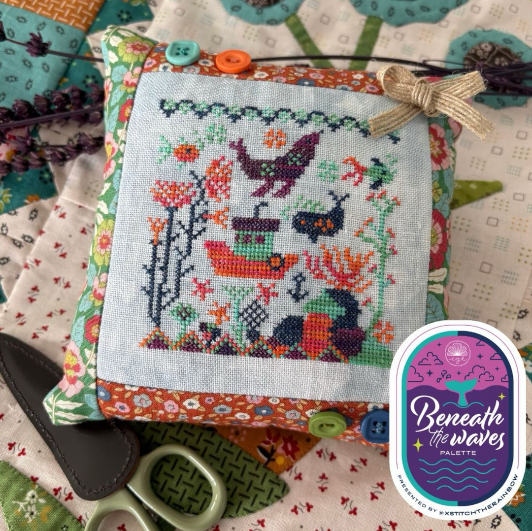 Cross-stitched pillow with colorful design on a fabric background, featuring the 'Beneath the Waves Palette' brand.