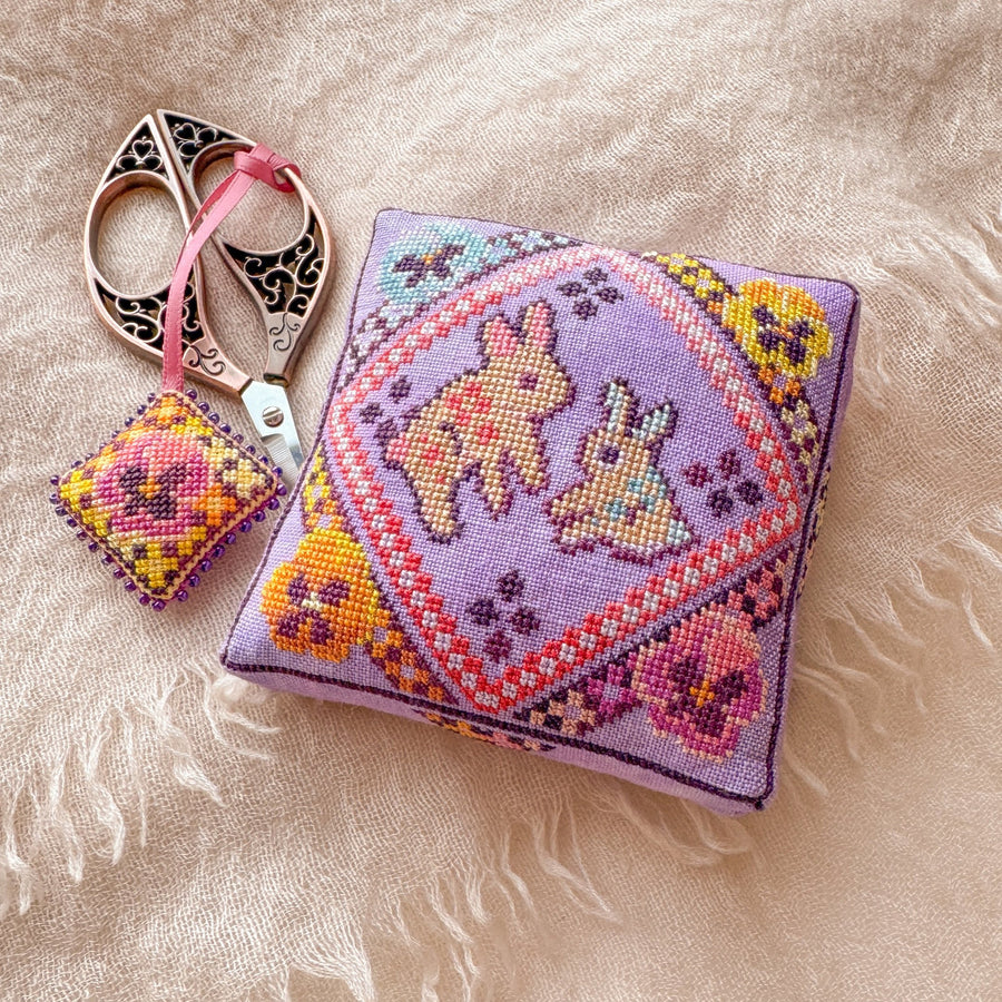 Cross-stitched pillow with two bunnies  and a small square pillow with floral pattern on a textured beige surface.