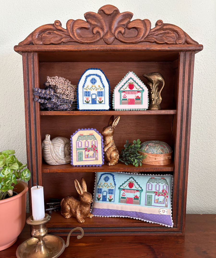 Wooden shelf with decorative items including cross-stitched house patterns, rabbits, and a candle.