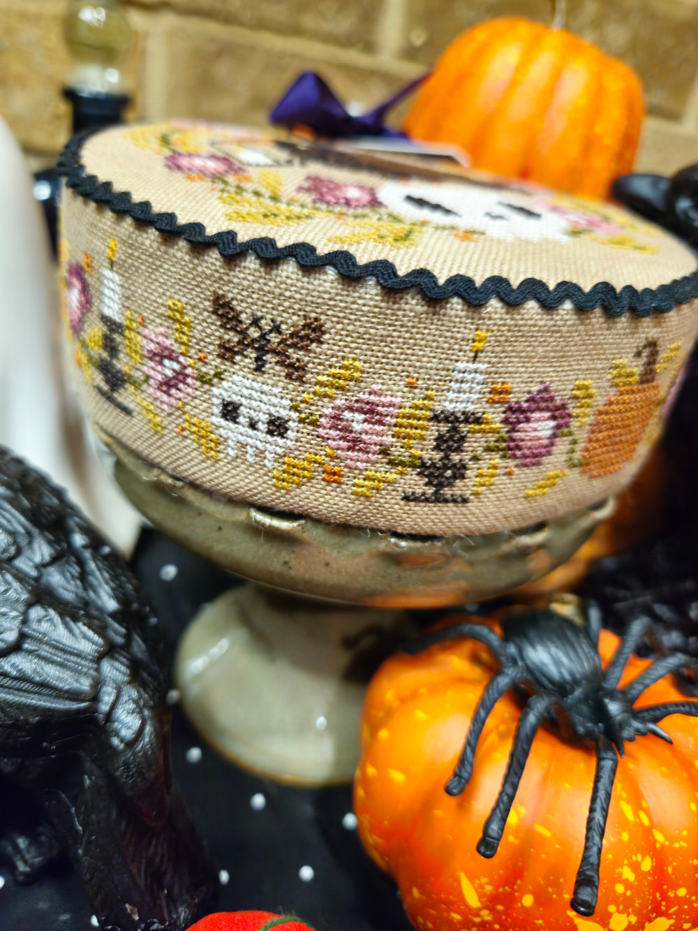 A close-up of a round cross-stitch sampler with a floral and skull pattern on beige linen, featuring a black scalloped edge and a small metal stand. The sampler is surrounded by Halloween decorations, including pumpkins and a black spider.