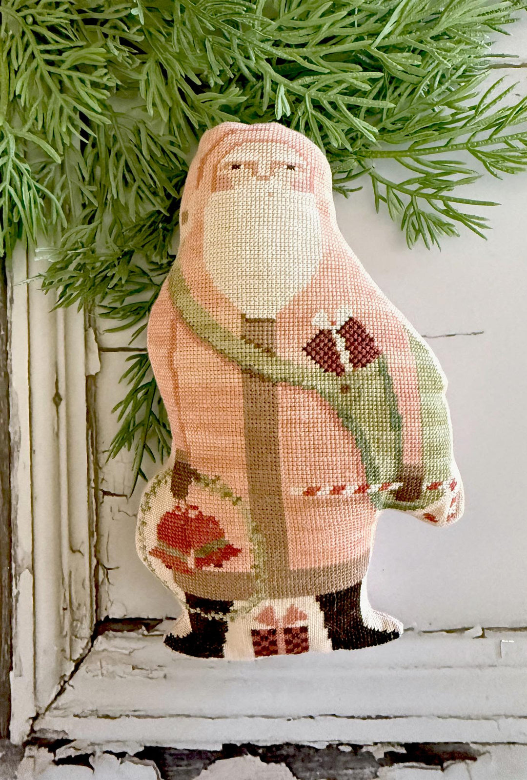 Decorative needlepoint of a santa wearing a  pink sweater with a green scarf, on a white surface with greenery.