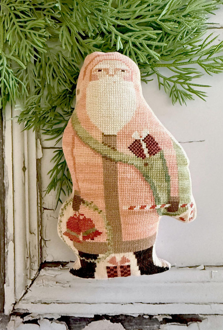 Decorative needlepoint of a santa wearing a  pink sweater with a green scarf, on a white surface with greenery.
