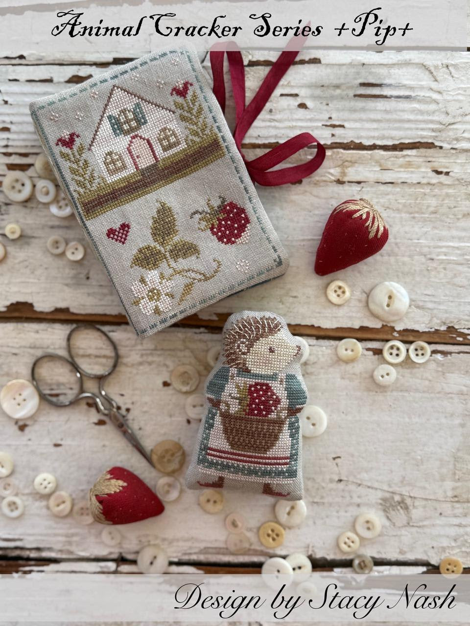 Cross-stitched design of a house and hedgehog with sewing tools on a wooden surface, featuring the Animal Cracker Series by Pip+. Design by Stacy Nash.