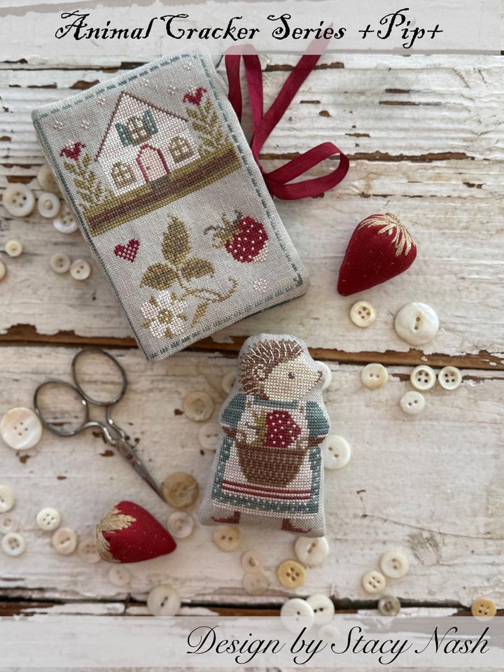 Cross-stitched design of a house and hedgehog with sewing tools on a wooden surface, featuring the Animal Cracker Series by Pip+. Design by Stacy Nash.