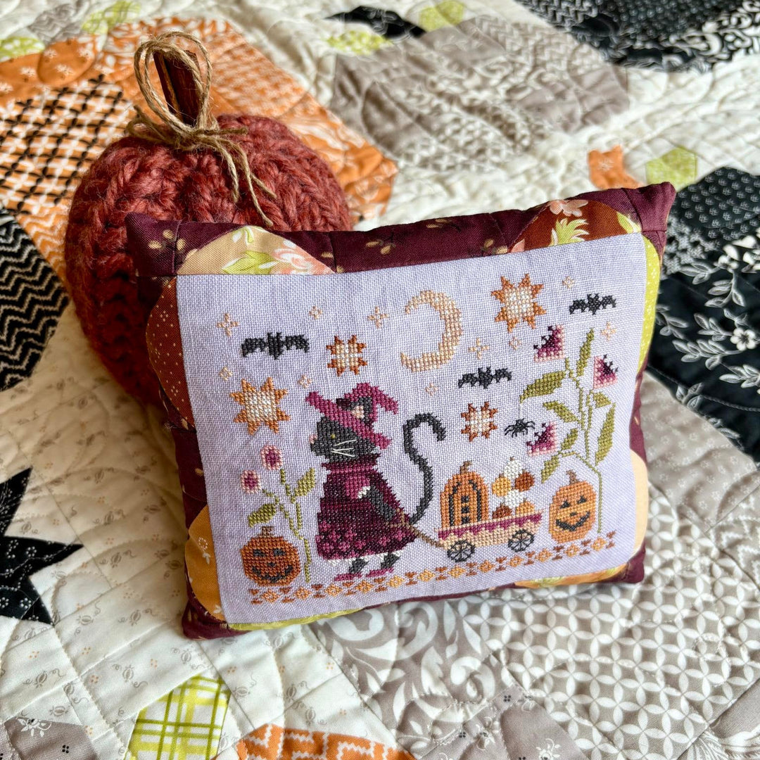 A small decorative pillow with a stitched Halloween design featuring a cat in a pumpkin carriage, surrounded by bats, flowers, and pumpkins, styled on a patchwork quilt next to a knitted pumpkin.