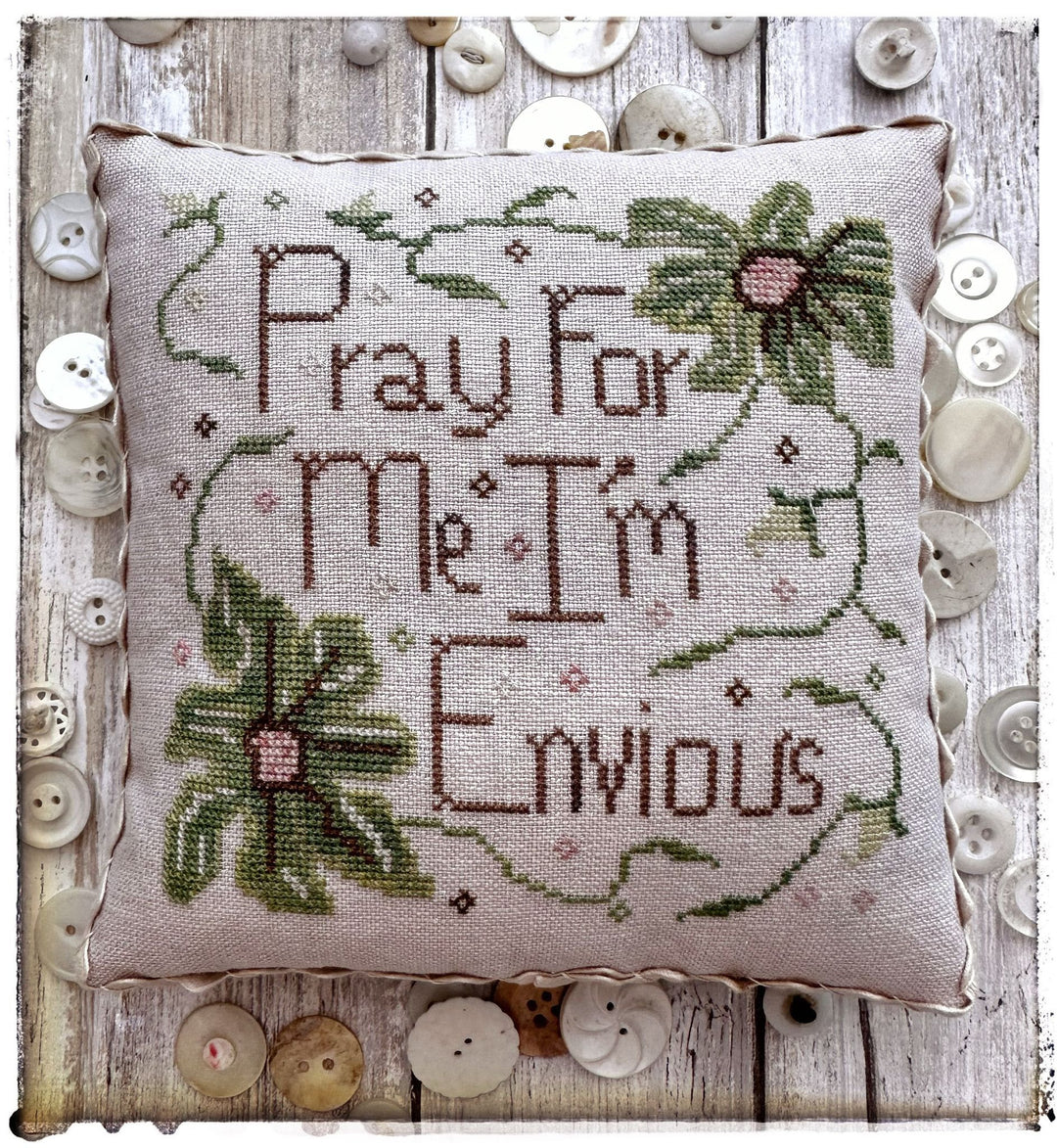 Decorative pillow with cross-stitched text and floral designs on a wooden surface with buttons.