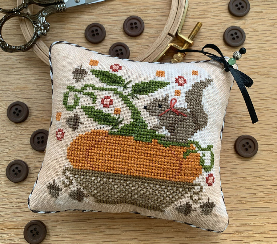 A square cross-stitch pillow with a squill on a pumpkin in a basket, placed on a wooden surface with embroidery hoops, scissors, and buttons around it.