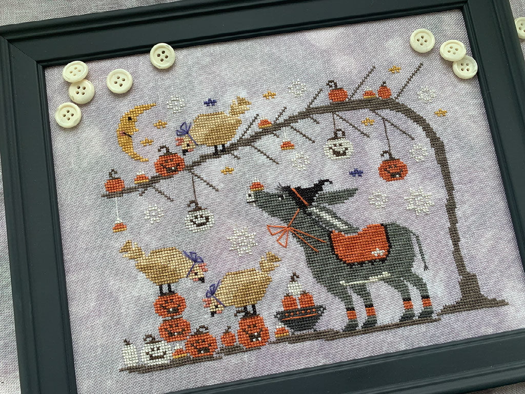 A framed cross-stitch artwork featuring a whimsical autumn scene with a gray donkey carrying an orange sack, pumpkins, hanging jack-o'-lanterns, birds, a crescent moon, and decorative buttons on the frame.