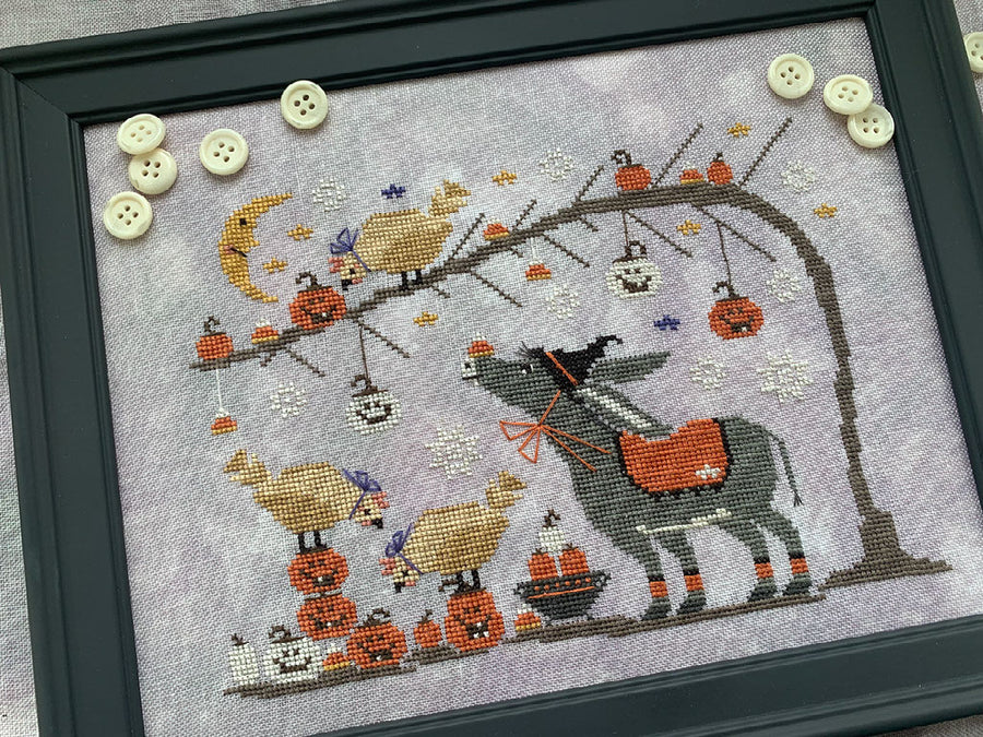 A framed cross-stitch artwork featuring a whimsical autumn scene with a gray donkey carrying an orange sack, pumpkins, hanging jack-o'-lanterns, birds, a crescent moon, and decorative buttons on the frame.