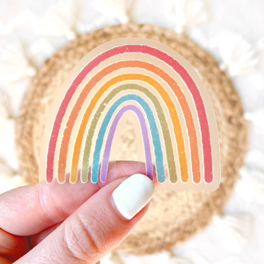 A hand holding a clear sticker with a colorful rainbow design. The sticker is intended to be applied to a surface to add a colorful rainbow motif.