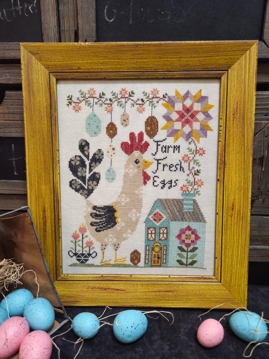 Framed cross-stitched picture with a rooster and house design, surrounded by Easter eggs.