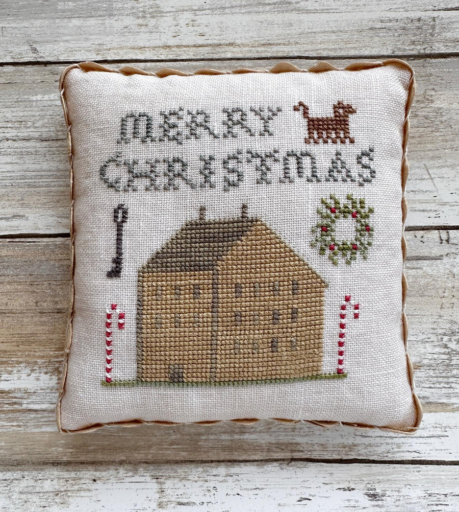 Decorative pillow with cross-stitched design of a house, candy cane, and deer on a wooden background.