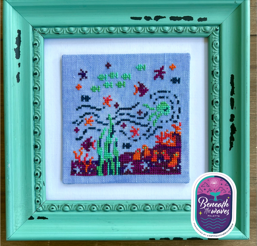 Framed cross-stitched artwork of an underwater scene with fish and coral, branded 'Beneath the Waves'.