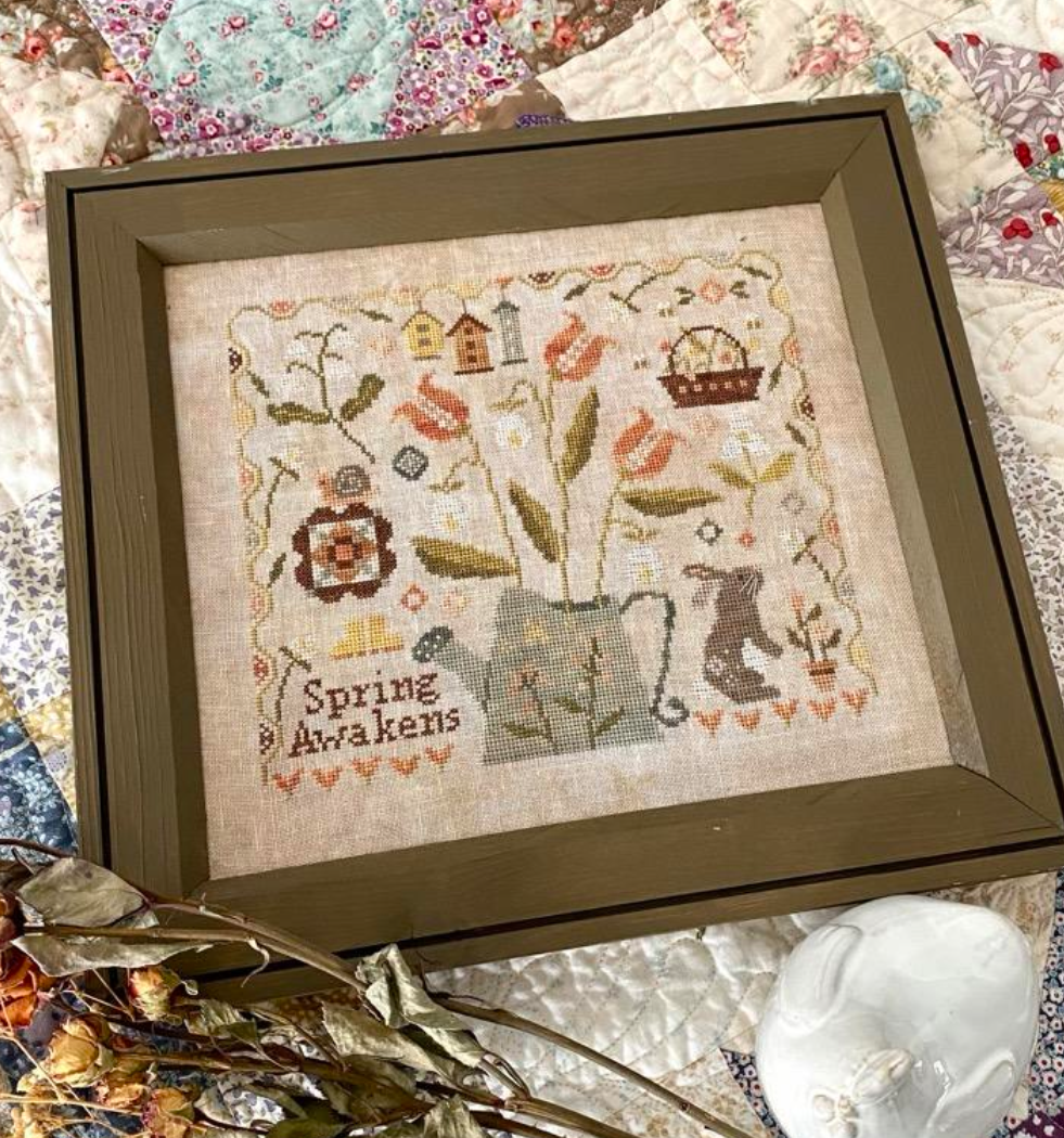 Framed cross-stitch art with 'Spring Awakens' text on a textured fabric background