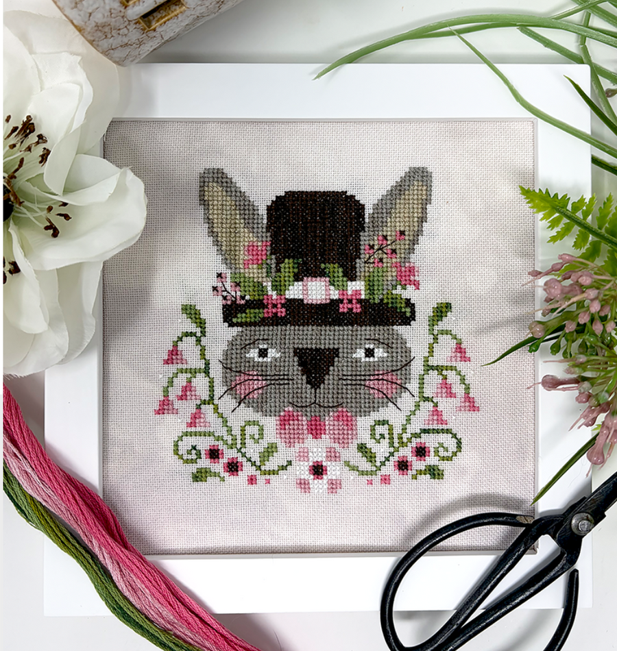 Cross-stitched bunny design with floral details on fabric, surrounded by flowers and scissors.