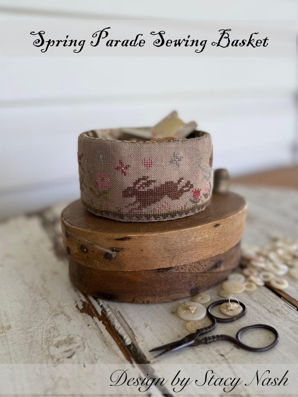 Sewing basket with a fabric roll and scissors on a wooden surface, labeled 'Spring Parade Sewing Basket' by Stacy Nash.