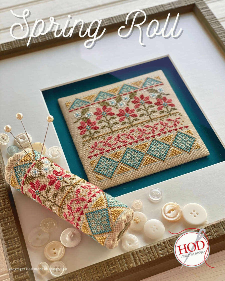 Framed cross-stitched piece with a spring roll design, surrounded by buttons and a pincushion.