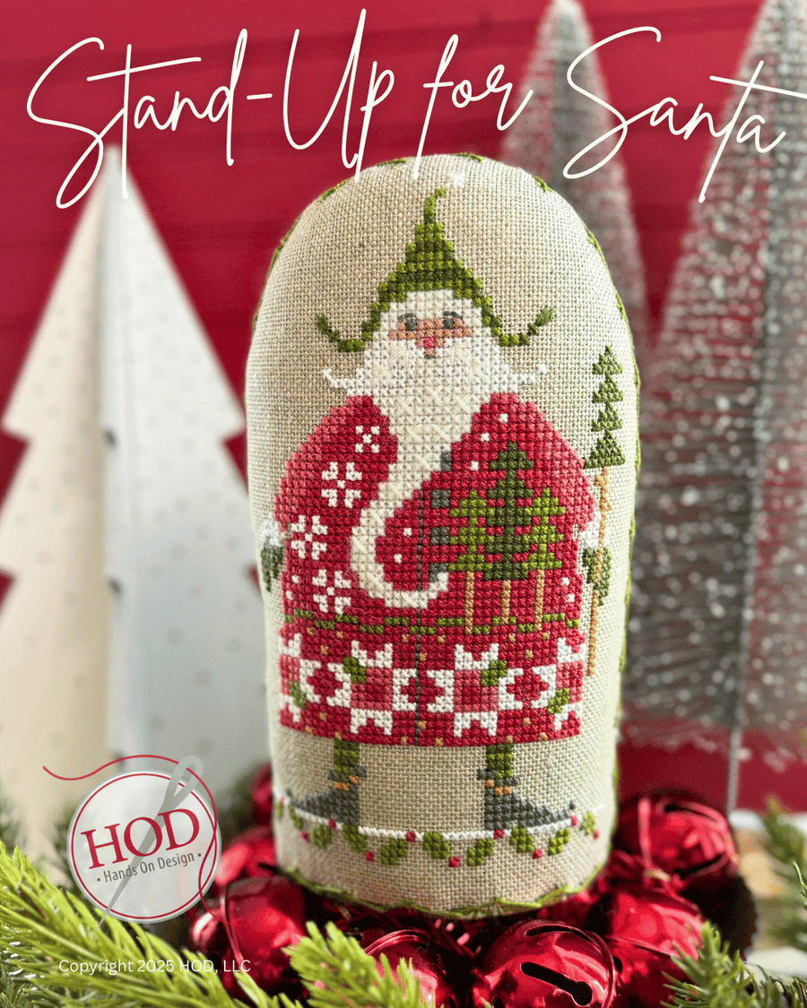 A cross stitch pillow featuring a festive Santa Claus design, displayed on a red and silver holiday backdrop with pine branches and red ornaments. The text 'Stand-Up for Santa' is written at the top, and a logo for 'HOD A Handy Oh Designs' is visible in the bottom left corner.