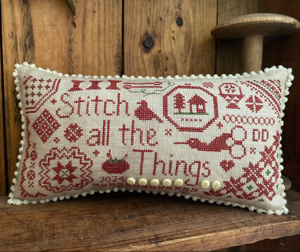 A rectangular cross-stitch pillow with a cream fabric base and red embroidered text reading 'Stitch all the Things,' plus decorative needlework motifs and a row of cream buttons along the bottom edge, displayed on a wooden surface against a wood-paneled background.