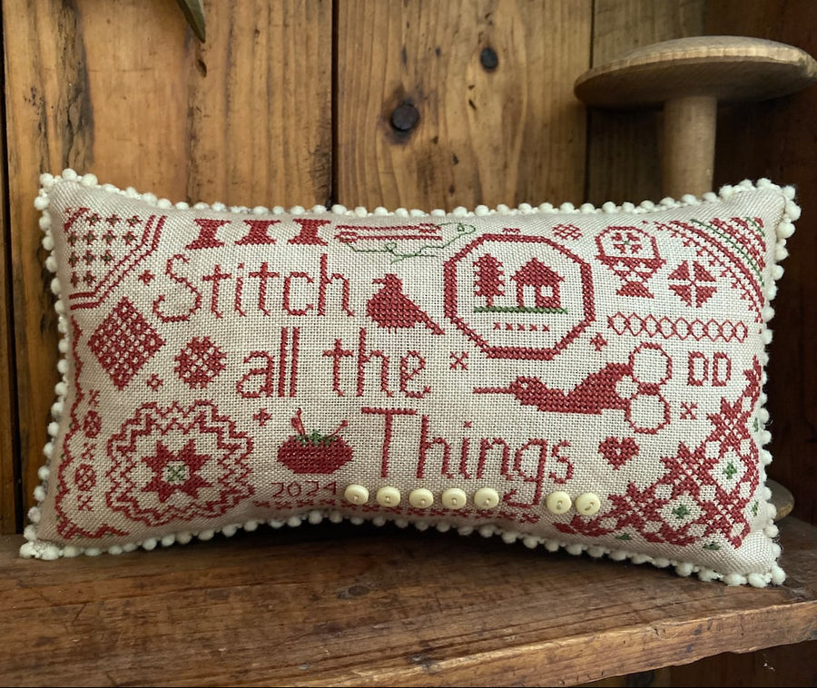 A rectangular cross-stitch pillow with a cream fabric base and red embroidered text reading 'Stitch all the Things,' plus decorative needlework motifs and a row of cream buttons along the bottom edge, displayed on a wooden surface against a wood-paneled background.