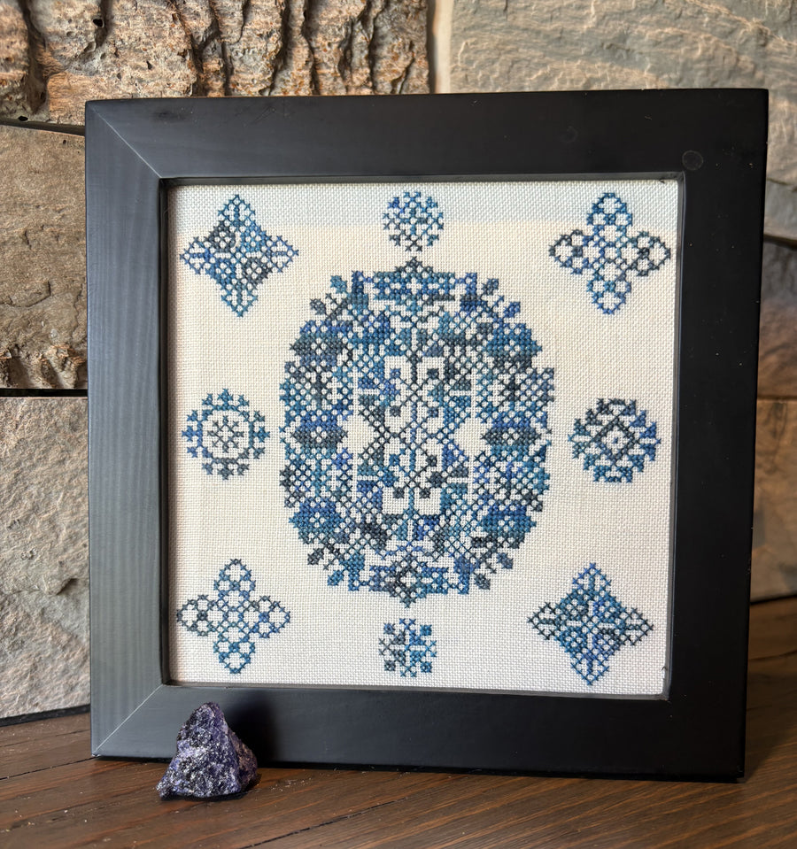 Framed cross-stitched pattern with blue geometric designs on a stone wall background