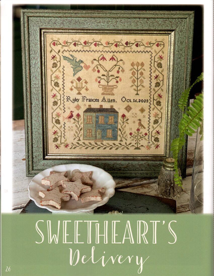 Framed cross-stitch pattern with a house design, placed on a surface with a plate of cookies and a plant.