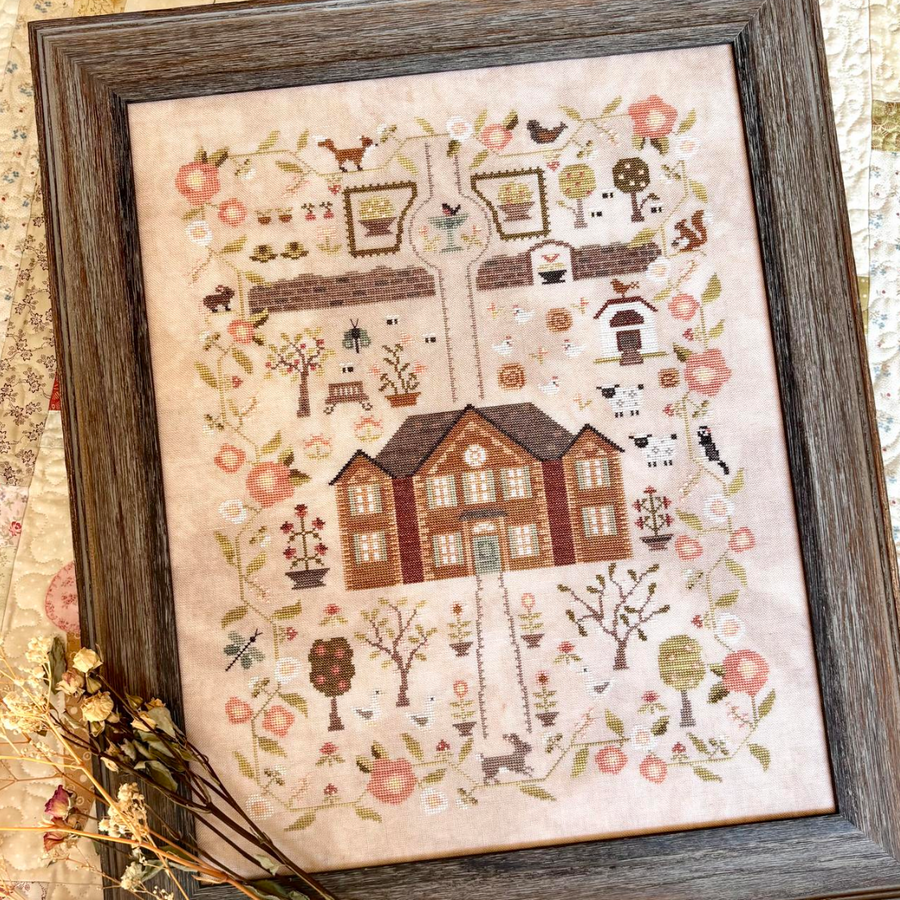 A framed cross-stitch artwork titled 'The Garden at Rosewood Hollow,' showing a detailed cottage scene with flowers, trees, birds, and small animals on a cream fabric background. The dark wood frame is shown with dried floral stems, and the title appears in a cream text panel below.