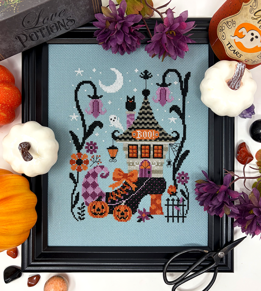 A framed cross-stitch artwork featuring a whimsical haunted house with pumpkins, bats, a crescent moon, and purple flowers, styled with small pumpkins, faux flowers, scissors, and a sign on a white background.