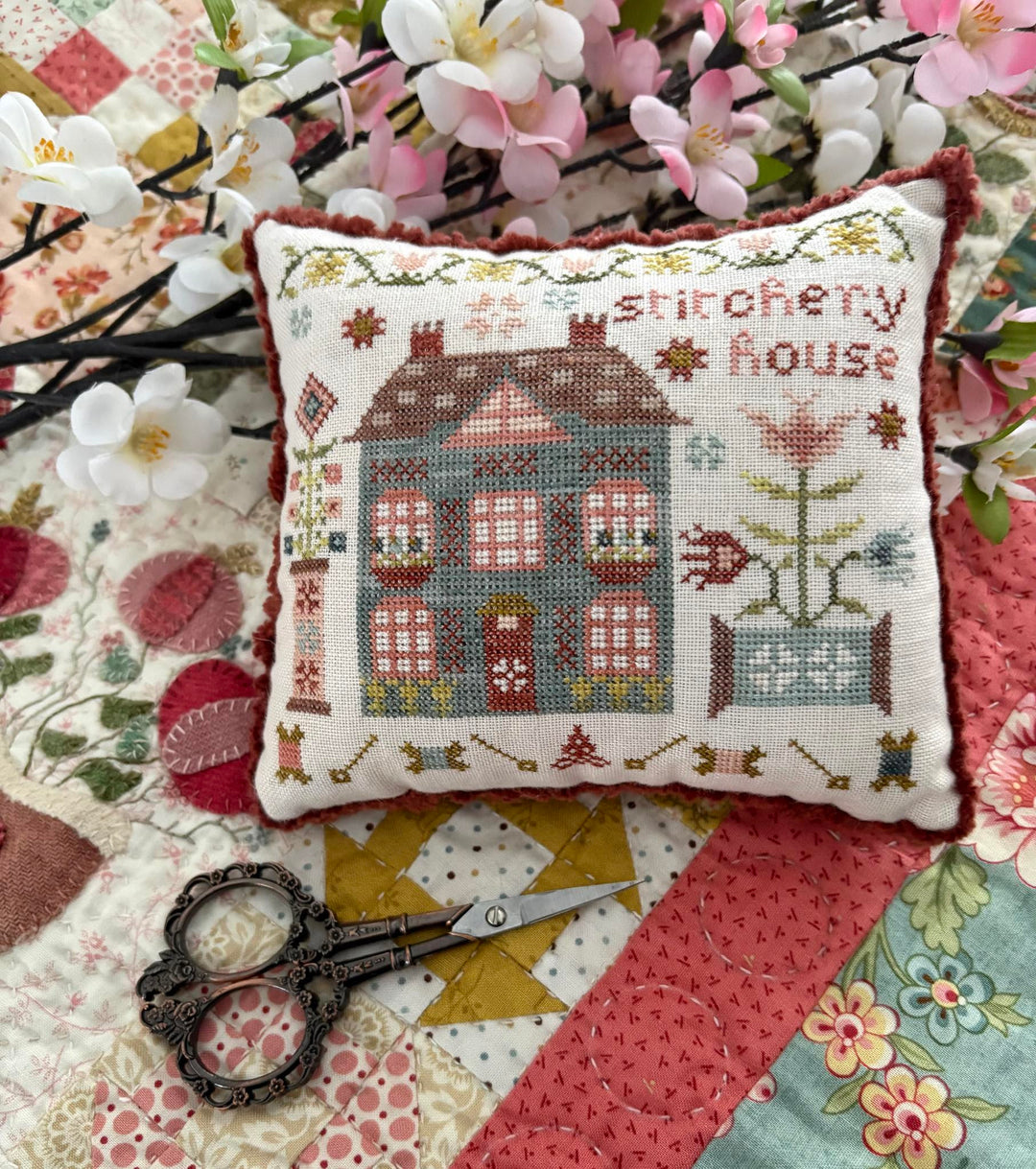 Decorative pillow with cross-stitched design of a house on a quilted fabric background.